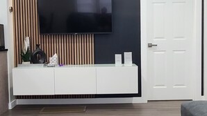 A modern living room with a flat-screen TV mounted on a wall with vertical wooden panels, a white entertainment unit below, and a glass coffee table in the foreground. - Contemporary 3 bedroom apartment on the first floor overlooking the Salmon River (Pulaski)
