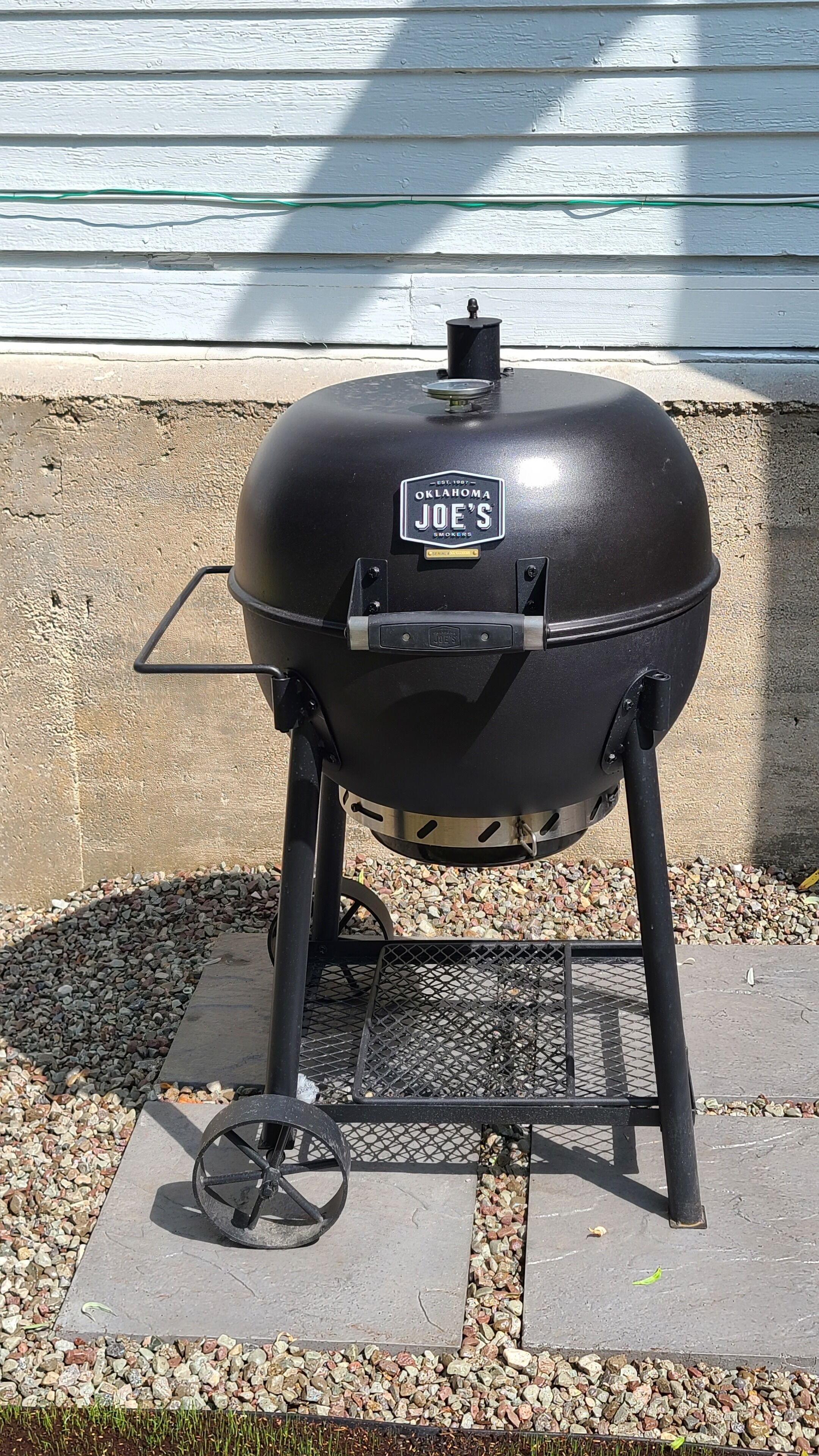 Un barbecue portable avec une étiquette indiquant « Oklahoma Joe's Smokers », posé sur une surface en béton, muni d'une grille en métal et de roulettes.