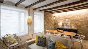 A cozy living room with a sofa, dining table, and chairs. There are large windows with sheer curtains, a stone wall, and exposed wooden beams on the ceiling.