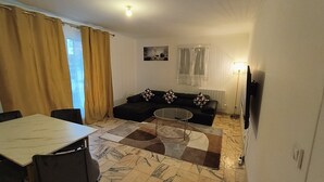 Ein Wohnzimmer mit einer Couch, einem Glastisch, einem Esstisch und einem Fernseher.