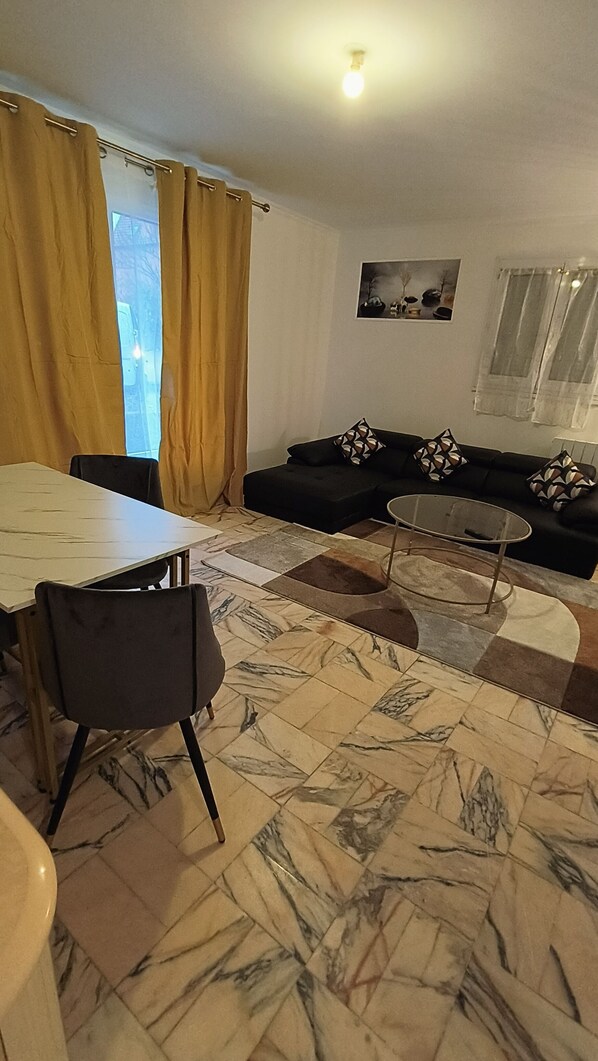 A living room with a sofa, a glass coffee table, a dining table with chairs, and a patterned rug.