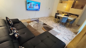 Ein Wohnzimmer mit einer schwarzen Couch, einem Glastisch und einem an der Wand befestigten Fernseher, der eine Nachrichtensendung zeigt.
