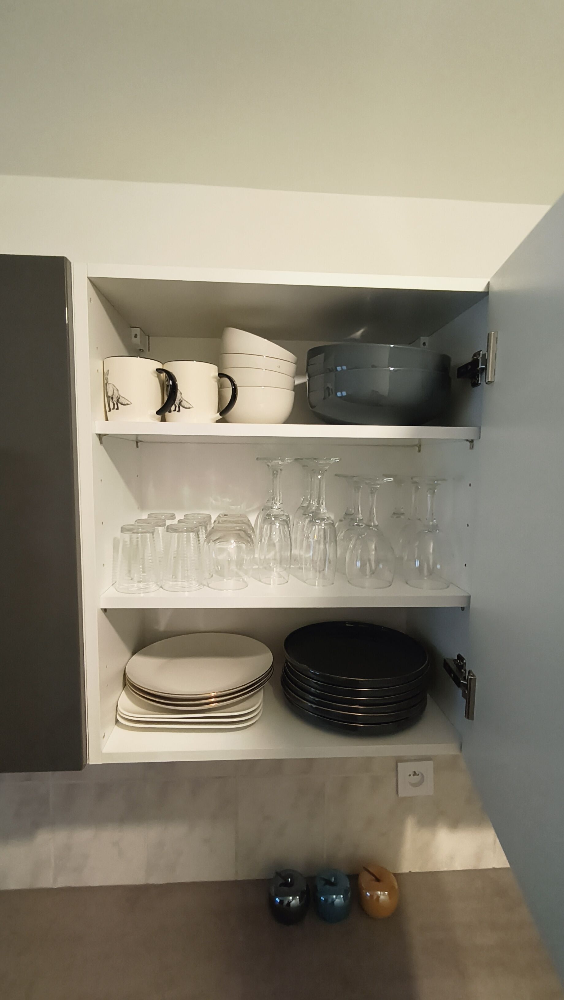 Ein Küchenschrank mit Geschirr, Gläsern und einem kleinen Gerät in Weiß und Schwarz.