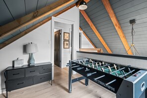 A room with a foosball table, a desk with drawers, a lamp, and a ceiling with wooden beams.