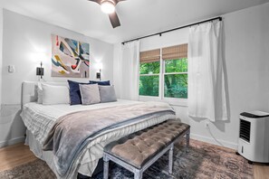 A bedroom with a bed, a bench, a window with curtains, a ceiling fan, and a wall-mounted air conditioner.