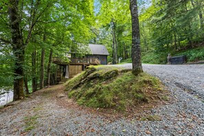 A cabin with a wooden deck and stairs leading to a door, surrounded by trees and a gravel path.