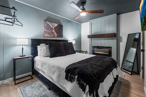 A modern bedroom with a bed, bedside tables, a ceiling fan, a fireplace, and a mirror.