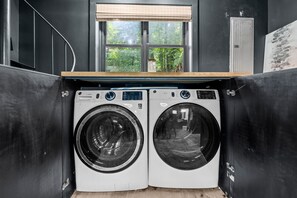 A compact laundry area with two front-loading washing machines, a window with blinds, and a wooden counter.