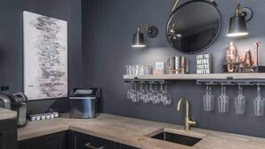 A modern kitchen with dark cabinetry, a wooden countertop, a sink, a round mirror, and a shelf with glasses and bottles.