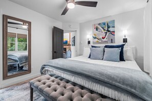 A bedroom with a large bed, a ceiling fan, a fireplace, and a view of the outdoors.