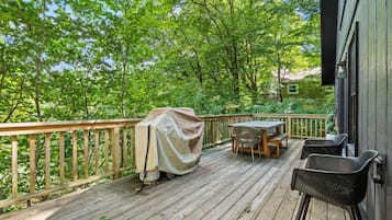 Een houten terras met een tafel, stoelen, een overdekte barbecue en uitzicht op een groen bos.