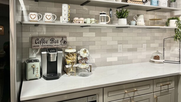Una cocina con baldosas blancas, un letrero de cafetería, una cafetera, un tostador y varios utensilios de cocina en estantes.