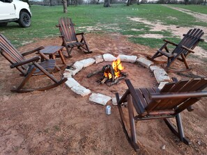 A circular arrangement of wooden chairs around a campfire in a dirt area with a white vehicle in the background.