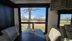 A travel trailer interior with a table and two chairs, a window with a view of trees and a clear sky.