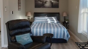 Een slaapkamer met een bed, een stoel, een plafondventilator, een schilderij en een raam met jaloezieën.