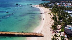 Une plage avec une jetée en bois, une eau turquoise et limpide, et une zone de villégiature agrémentée de palmiers et de chaises de plage.
