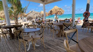 Une terrasse en bois, agrémentée de tables et de chaises, qui offre une vue sur une plage bordée de parasols en chaume et de palmiers.