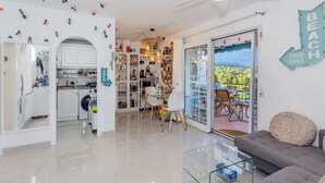 A modern living room with a sofa, a glass coffee table, and a dining area with chairs and a table. There is a kitchen area with cabinets and appliances, and a balcony with a view of greenery. - Flat Surrounded by Nature, With Pool, and Beach Nearby (Son Parc)