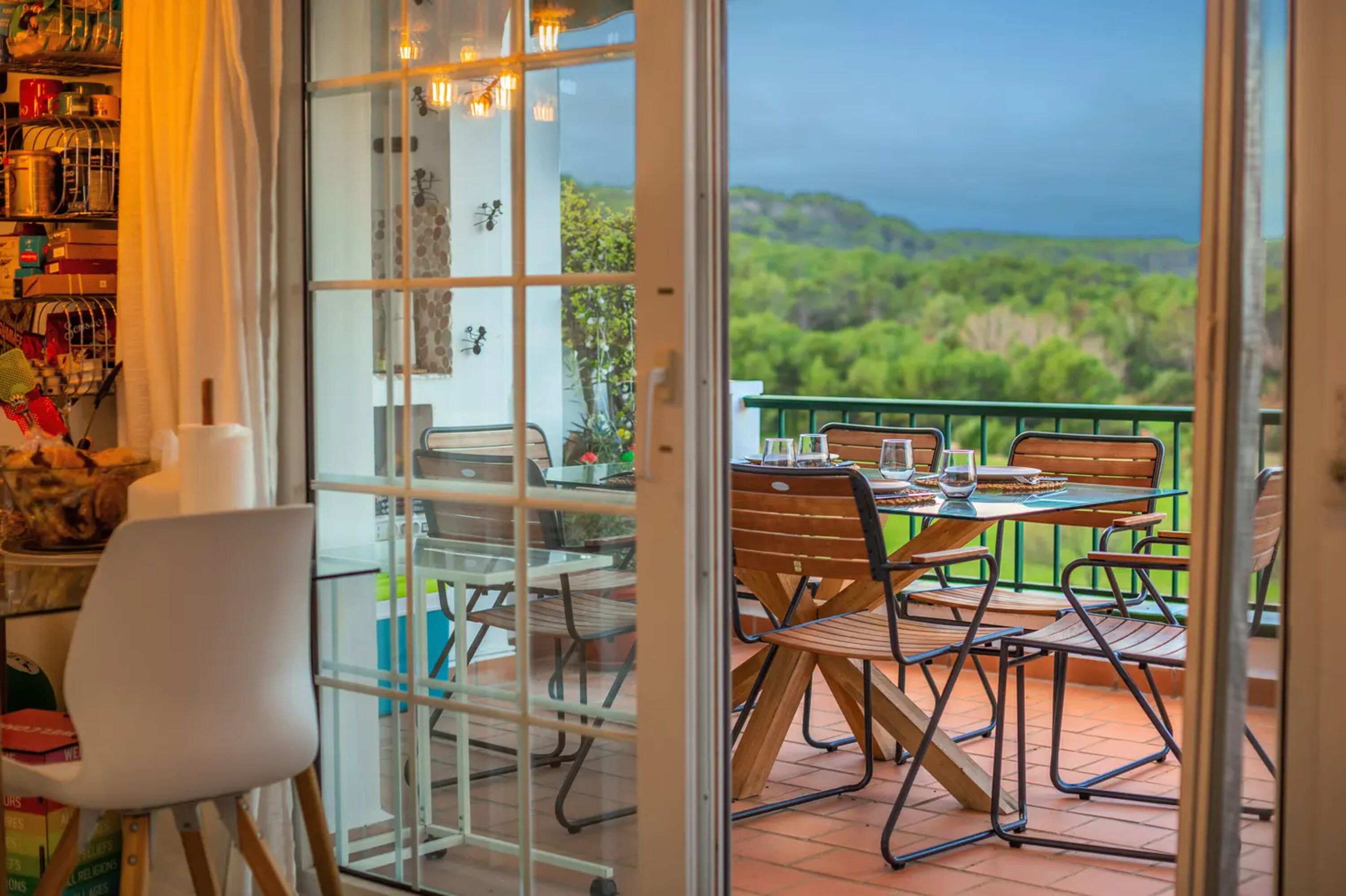 A balcony with a table and chairs, a view of greenery, and a glass door leading to a room with a bookshelf and a chair.