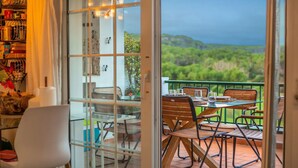 A balcony with a table and chairs, a view of greenery, and a glass door leading to a room with a bookshelf and a chair. - Flat Surrounded by Nature, With Pool, and Beach Nearby (Son Parc)