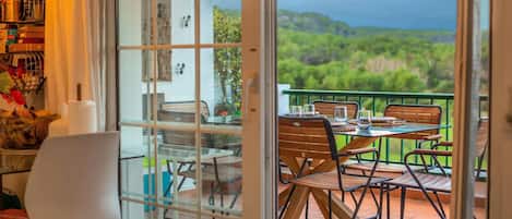 A balcony with a table and chairs, a view of greenery, and a glass door leading to a room with a bookshelf and a chair.