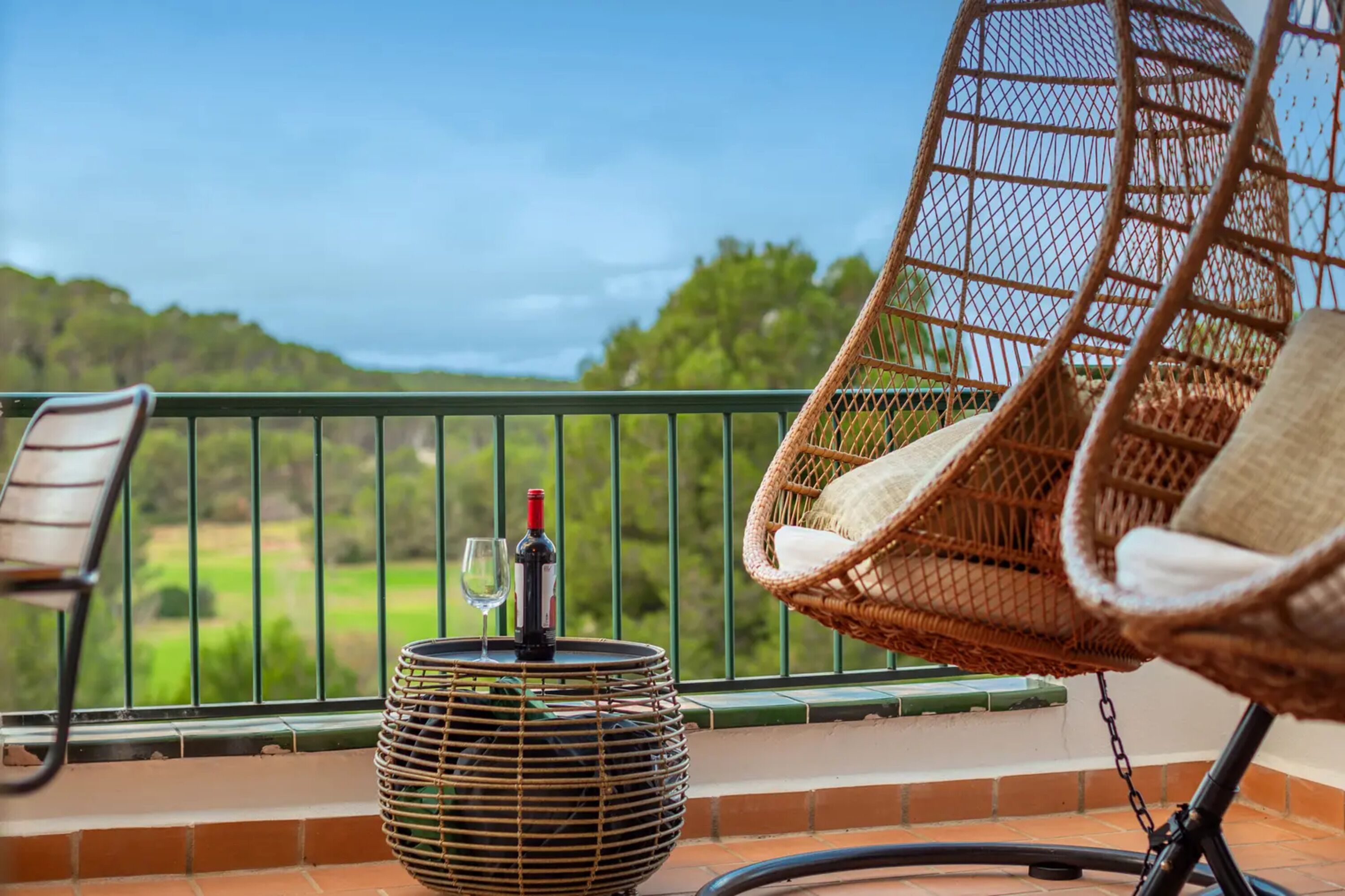 Un balcon avec des fauteuils en osier, une petite table avec une bouteille et un verre, et une vue sur un paysage verdoyant.