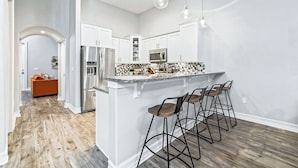 A modern kitchen with white cabinets, a stainless steel refrigerator, a microwave, and a granite countertop. There are three bar stools and pendant lights above the counter.