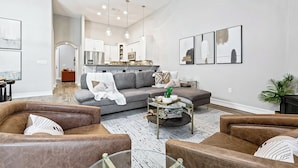 A spacious living room with a grey sofa, brown leather armchairs, and a coffee table. A kitchen is visible in the background with white cabinets and stainless steel appliances.