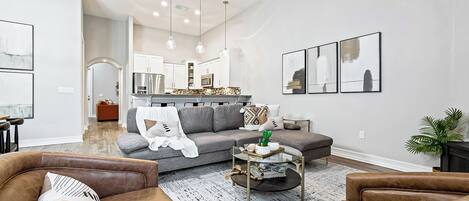 A spacious living room with a grey sofa, brown leather armchairs, and a coffee table. A kitchen is visible in the background with white cabinets and stainless steel appliances.