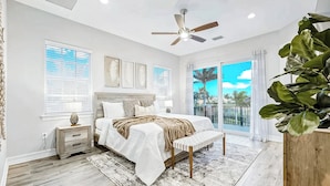A bedroom with a bed, bedside tables, a ceiling fan, and a large window with a view of palm trees.
