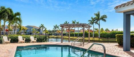 A shared community pool with a clear blue water, surrounded by a brick patio, palm trees, and a pergola.