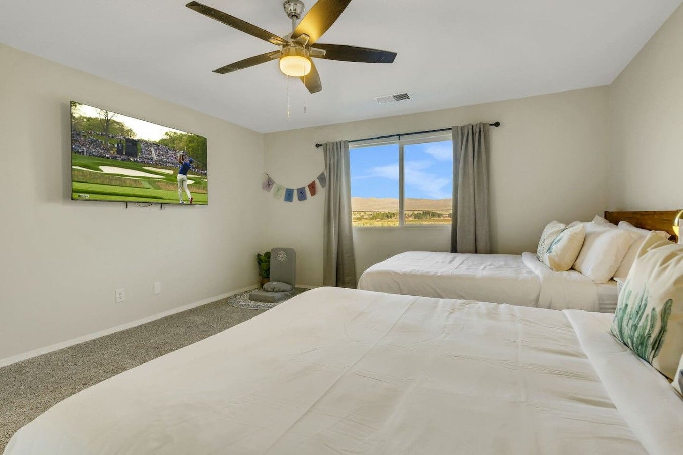 A bedroom with two beds, a ceiling fan, a large window with curtains, and a television on the wall.
