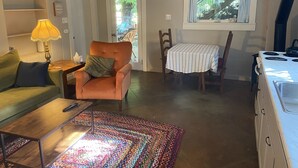 A cozy living room with a sofa, armchair, coffee table, and a colorful rug. There's a dining area with a table and chairs, and a kitchen with a stove.