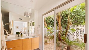 Un baño moderno con un espejo grande, armarios de madera y vistas a un jardín a través de una puerta de cristal.