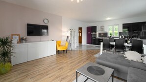 Un salon moderne avec un canapé gris, une table basse et un fauteuil jaune. On y trouve également un coin cuisine avec des meubles noirs et une table à manger. Un téléviseur à écran plat est fixé au mur, et une horloge est visible.