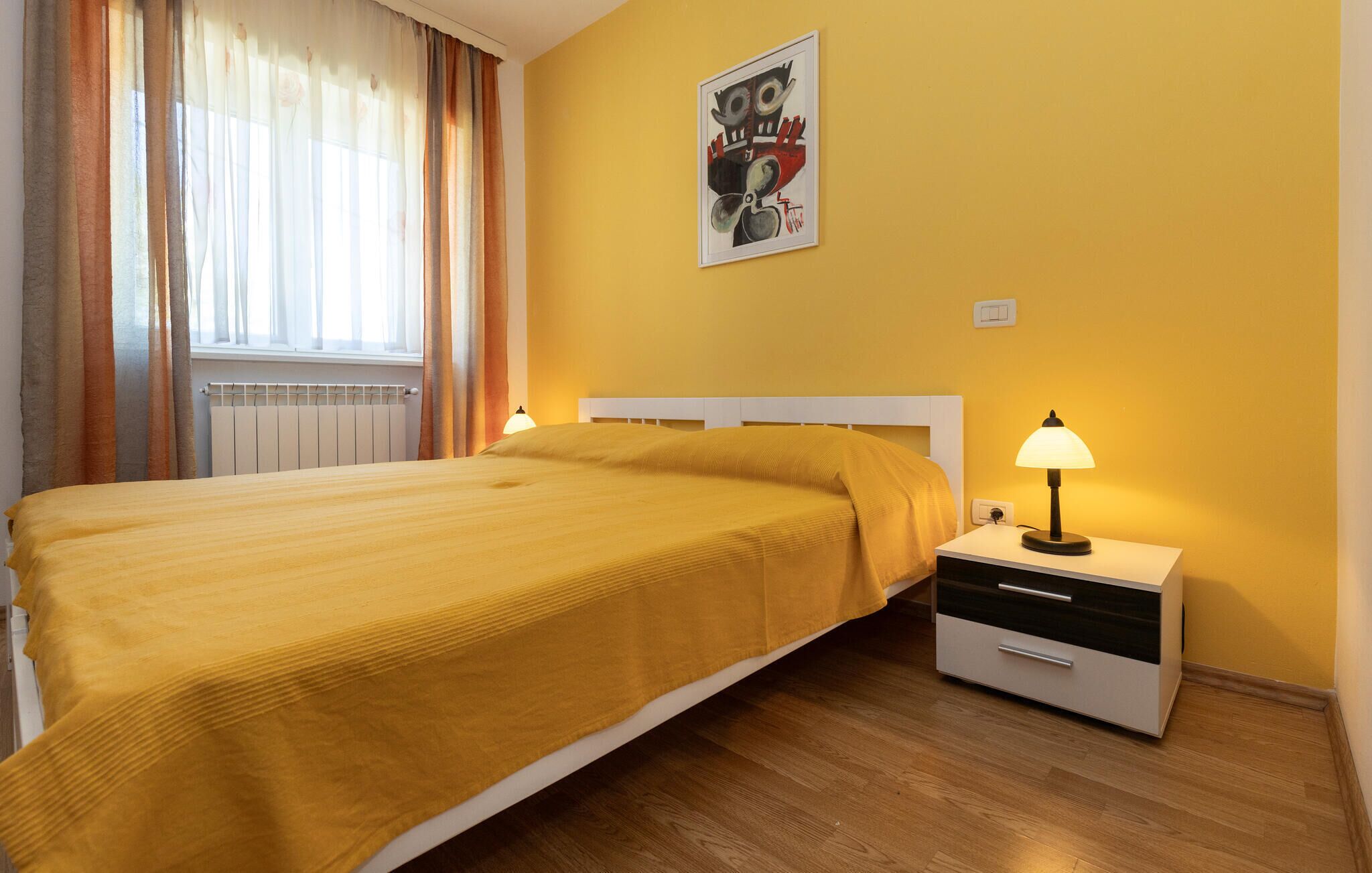 Une chambre avec un lit, une table de chevet, une lampe, une affiche au mur et une fenêtre avec des rideaux.