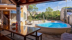 Zona de piscina exterior con mesa y sillas de madera, rodeada de muros de piedra y follaje.