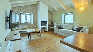 Ein gemütliches Wohnzimmer mit Steinwand, Holzbalken an der Decke, einem Flachbildfernseher, einem weißen Sofa und einem Holztisch.