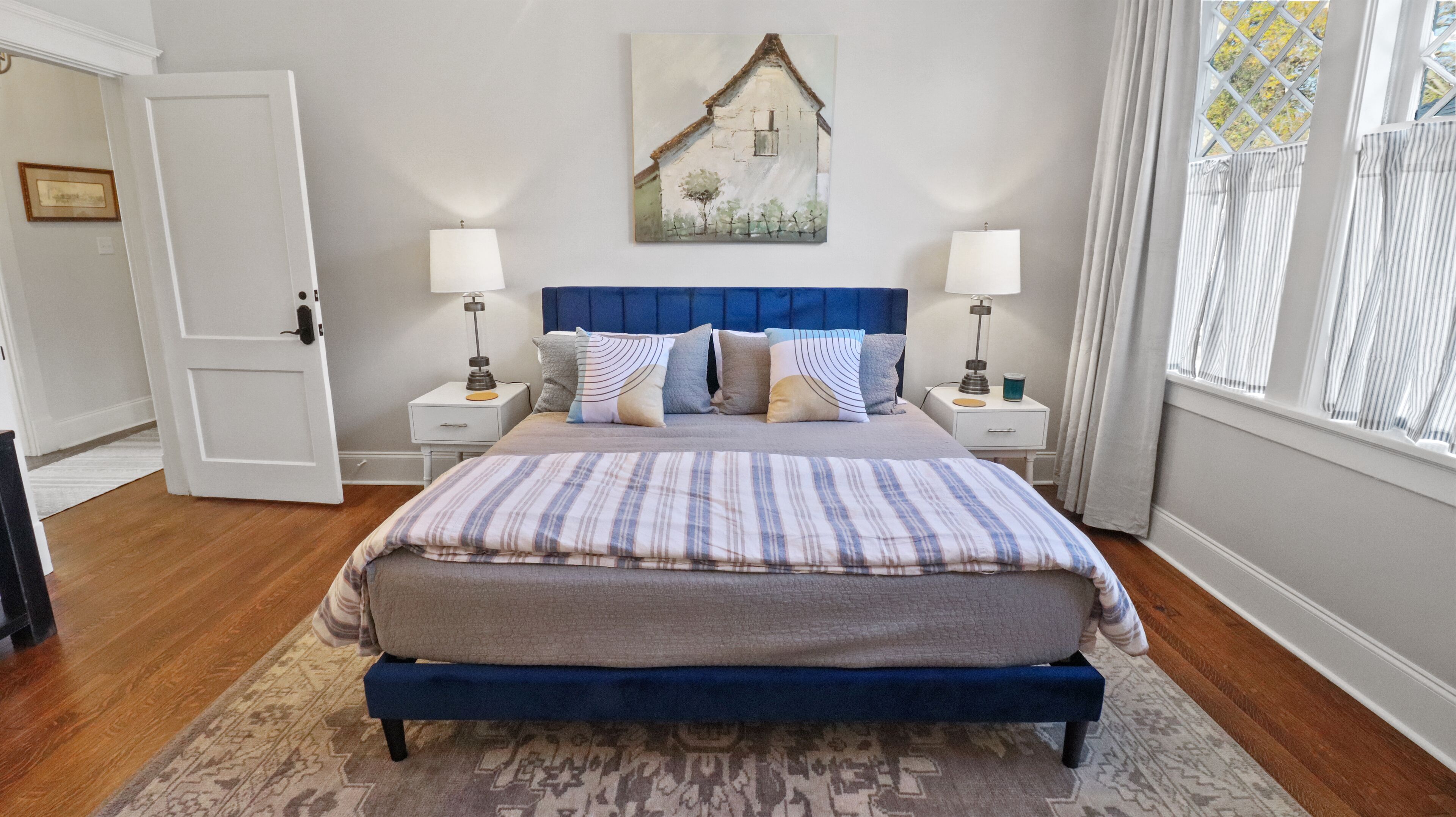 A bedroom with a bed, two bedside tables, a nightstand, a lamp, a painting on the wall, and a window with curtains.
