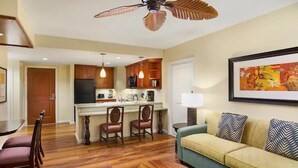 A living room with a green sofa, wooden dining table, and chairs. There is a kitchen area with wooden cabinets and a ceiling fan. - Waikiki | Waikiki Lagoon Escape | Pool & Beach | 2BR (Honolulu)