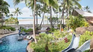 A pool with a slide, surrounded by palm trees and a beach view. - Waikiki | Waikiki Lagoon Escape | Pool & Beach | 2BR (Honolulu)