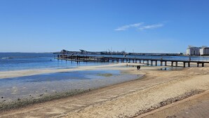 A beach with a pier extending into the water, a sandy shore, and industrial buildings in the background.