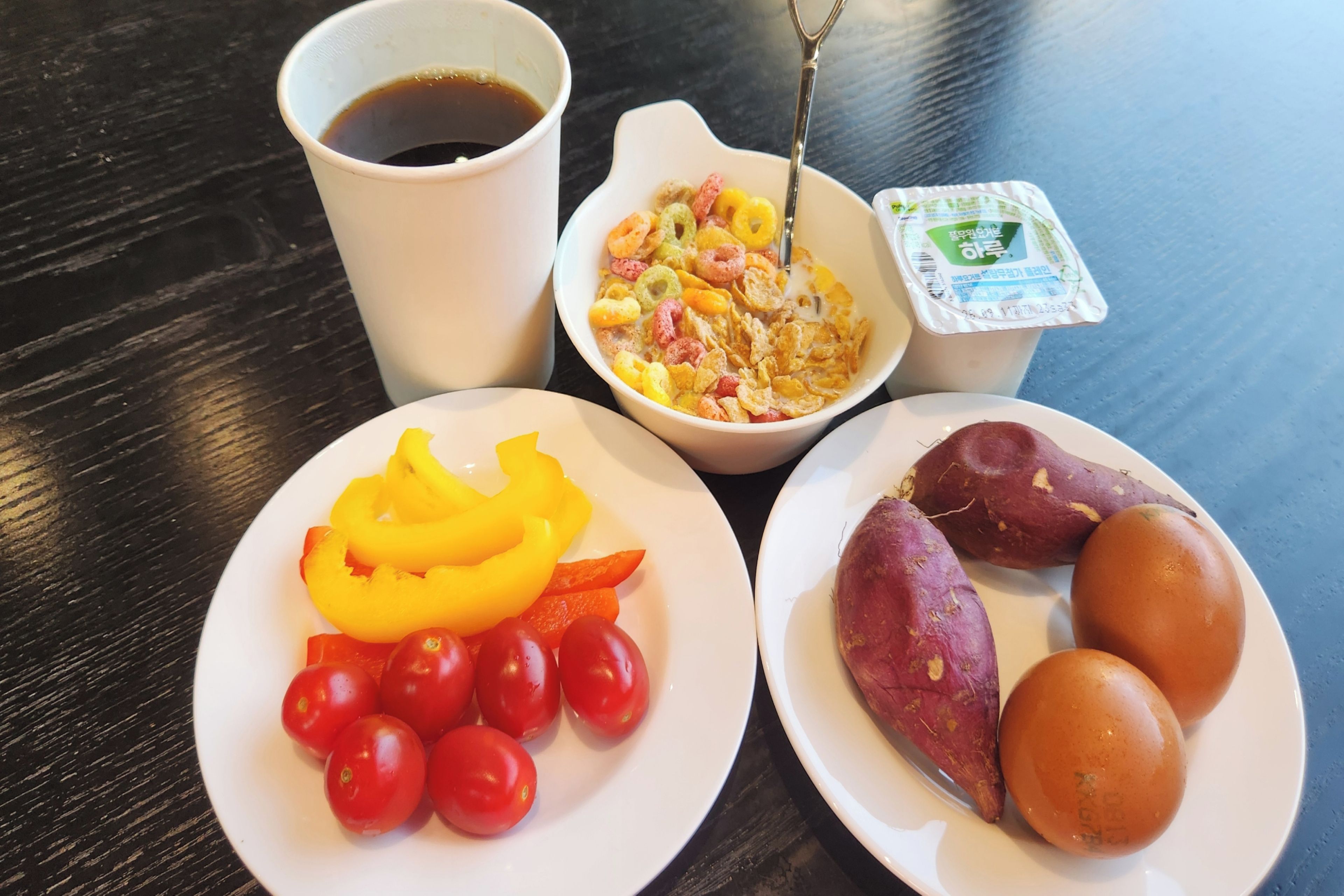 A table with a white plate of cherry tomatoes, a bowl of cereal, a cup of coffee, and a plate of sweet potatoes.