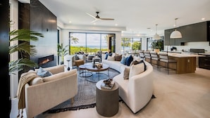A modern living room with a curved sofa, a coffee table, and a TV. There is a kitchen area with a bar and large windows offering a view of the outdoors. - Pier Pleasure - Anna Maria at it's finest! Pool, hot tub, beach, putting green (Anna Maria)