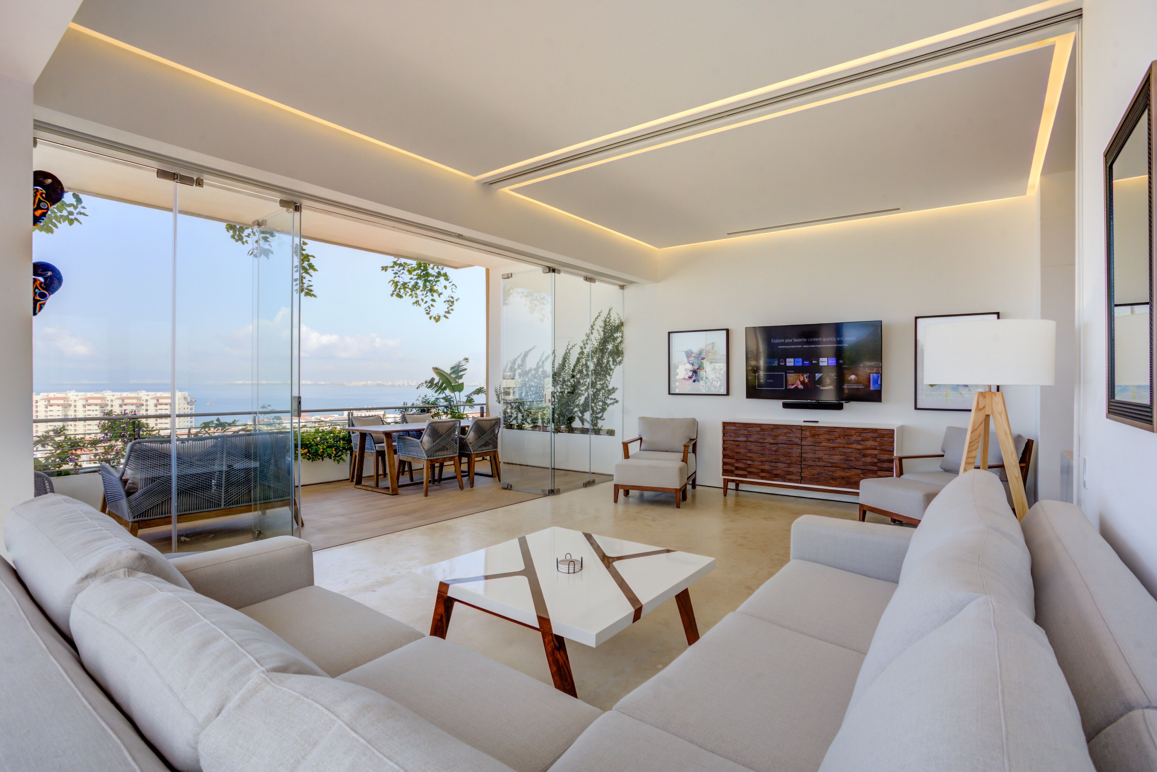 Un salón moderno con un sofá grande, una mesa de centro y un televisor. Tiene un balcón con vistas a la ciudad y al mar.