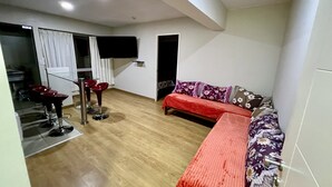 A compact living space with a sofa, a TV mounted on the wall, and a dining area with red chairs. - Acogedor Departamento en Lima - Jesús María, Ideal Para Familias (Jesús María)