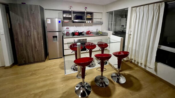 A modern kitchen with a refrigerator, microwave, and stove. There are four red bar stools in front of the counter. - Acogedor Departamento en Lima - Jesús María, Ideal Para Familias (Jesús María)