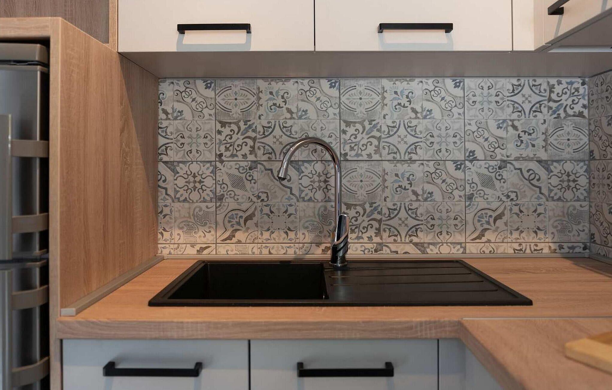 A kitchen with a black sink, patterned backsplash, wooden cabinets, and a stainless steel range hood.