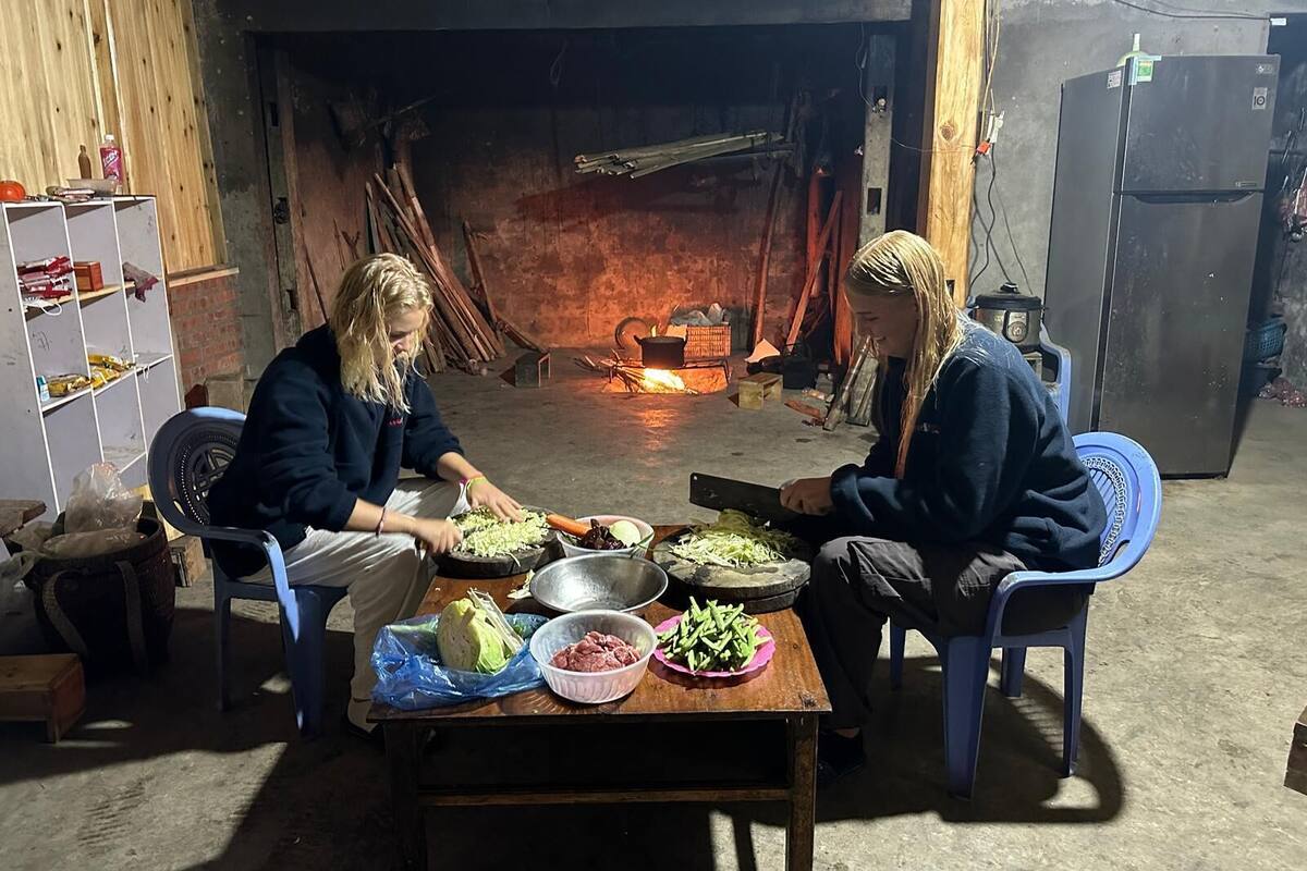 Two people preparing food at a wooden table with various vegetables and meat in a rustic room with a fireplace.