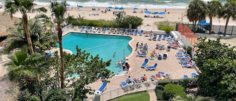 A pool area with lounge chairs and umbrellas, surrounded by palm trees and a view of the ocean.